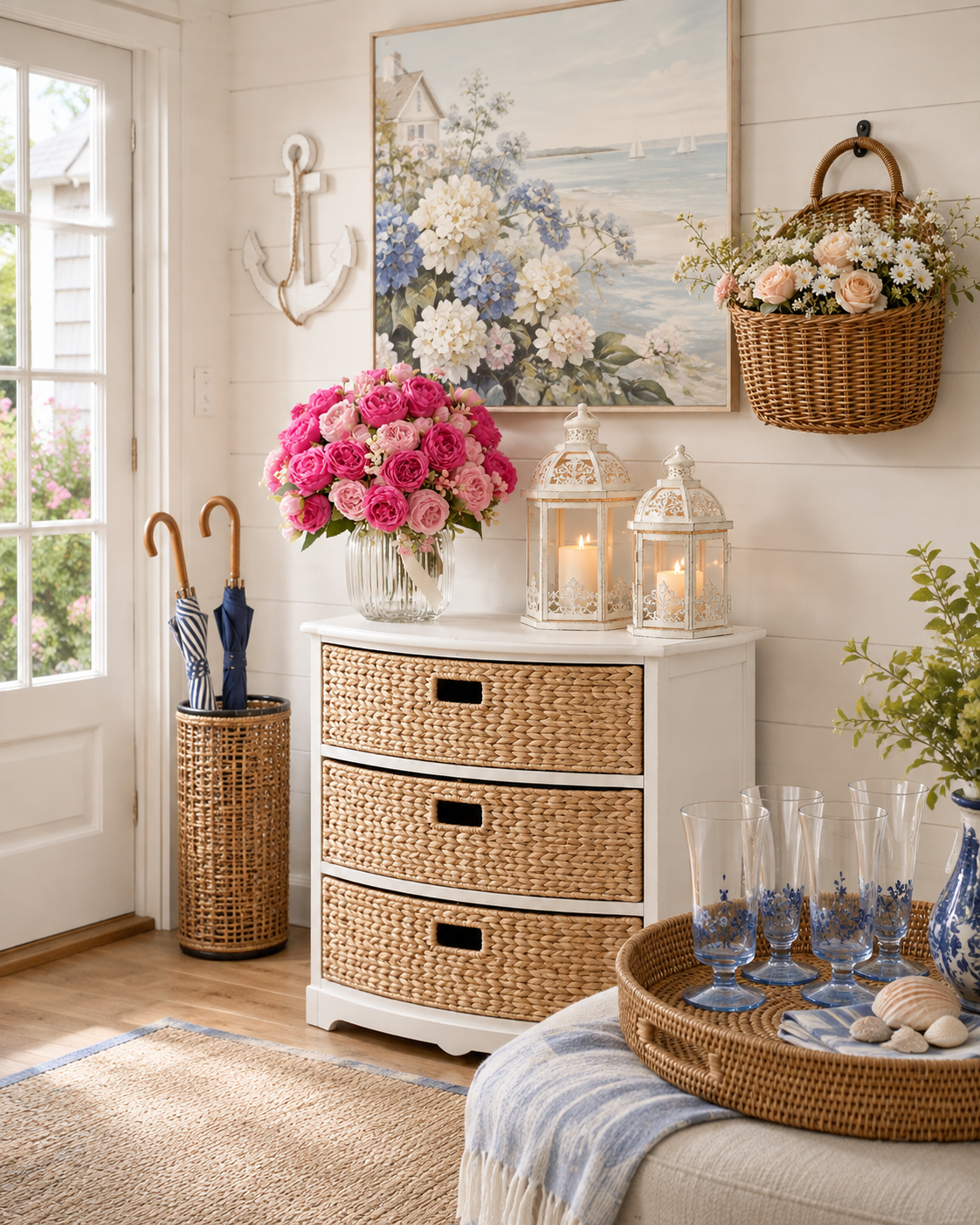 A pretty coastal entryway with a white curved cabinet featuring three woven water-hyacinth drawers, a large hydrangea-and-beach-house canvas above, a glass vase of pink roses, two white scroll lanterns, a woven wall basket filled with flowers, a distressed white anchor wall decor, a rattan umbrella stand, and a woven tray holding blue-stemmed goblets, seashells and a blue-and-white ginger jar.